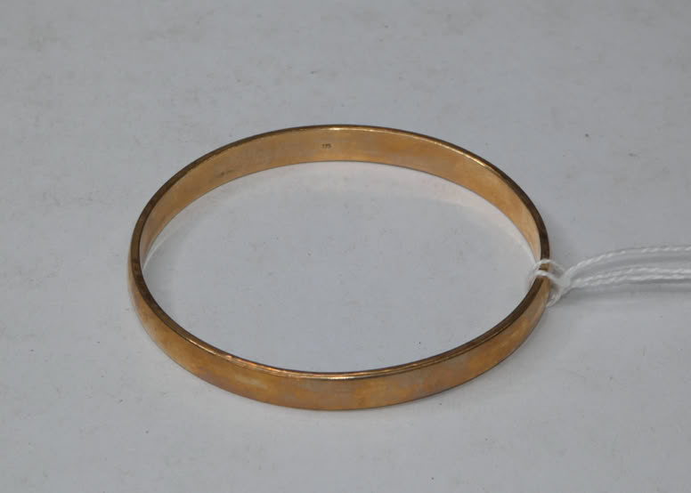 A gold-colored circular bracelet with a small white tag attached, placed on a light gray surface.