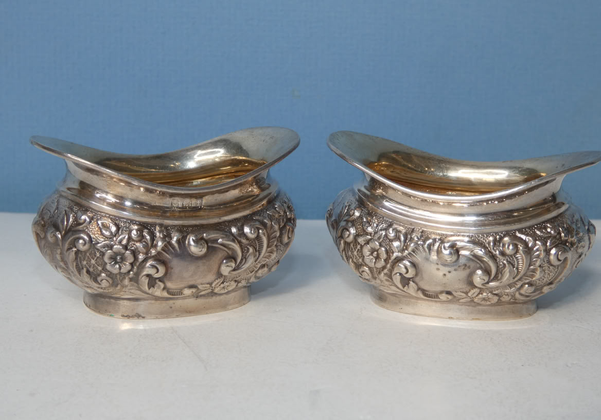 Two ornate silver gravy boats with floral engravings on a white surface against a blue background.
