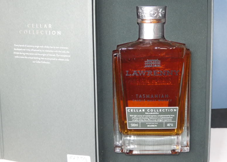 A bottle of amber-colored liquor in a rectangular glass bottle with a silver cap, placed inside an open box with text on the left side.