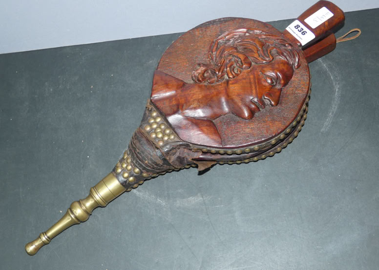 A decorative bellows with a carved wooden handle featuring a relief of a face, and a brass nozzle, resting on a dark surface.